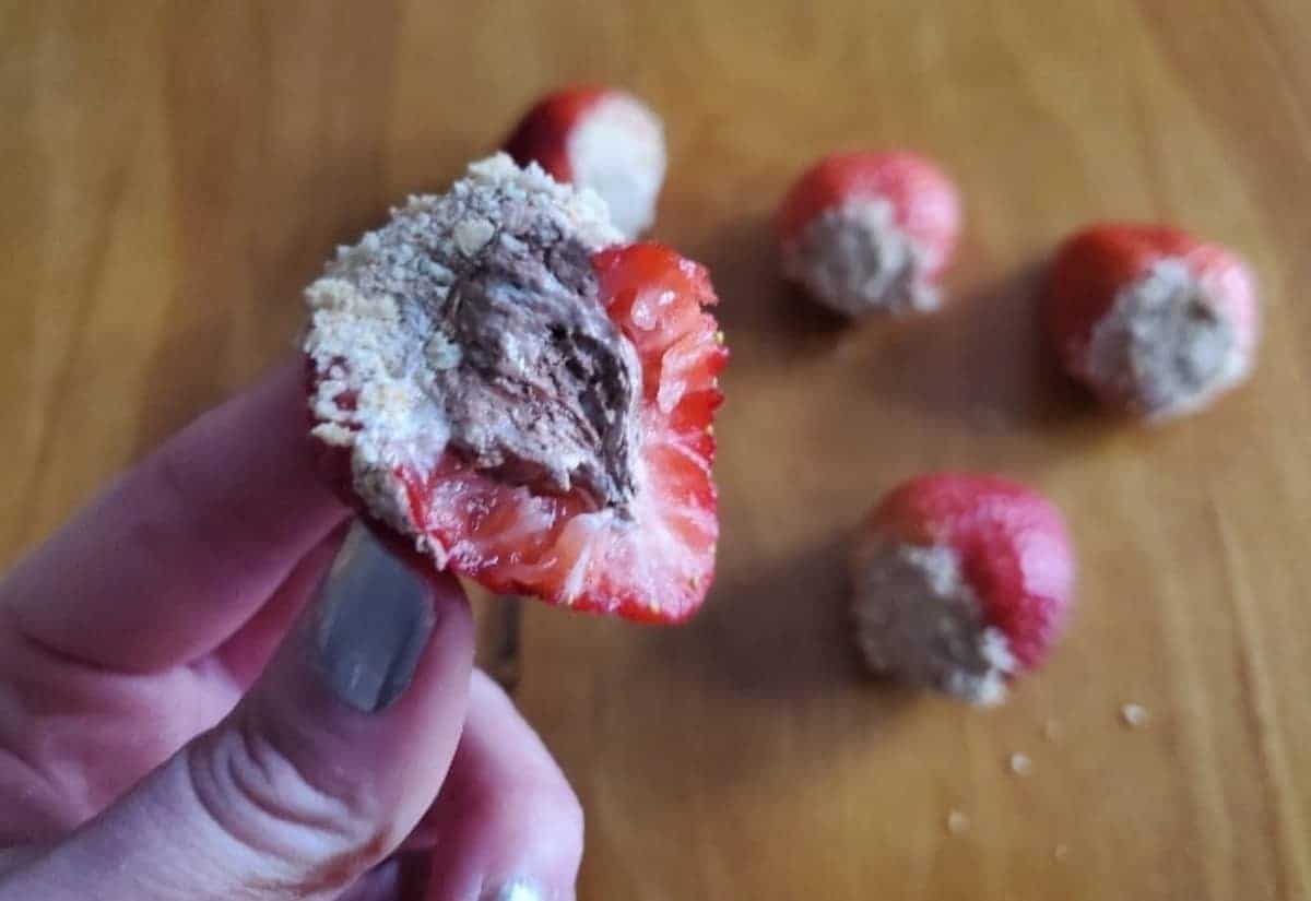 Image shows a hand holding a s'mores stuffed strawberry half with the filling exposed and more behind it.