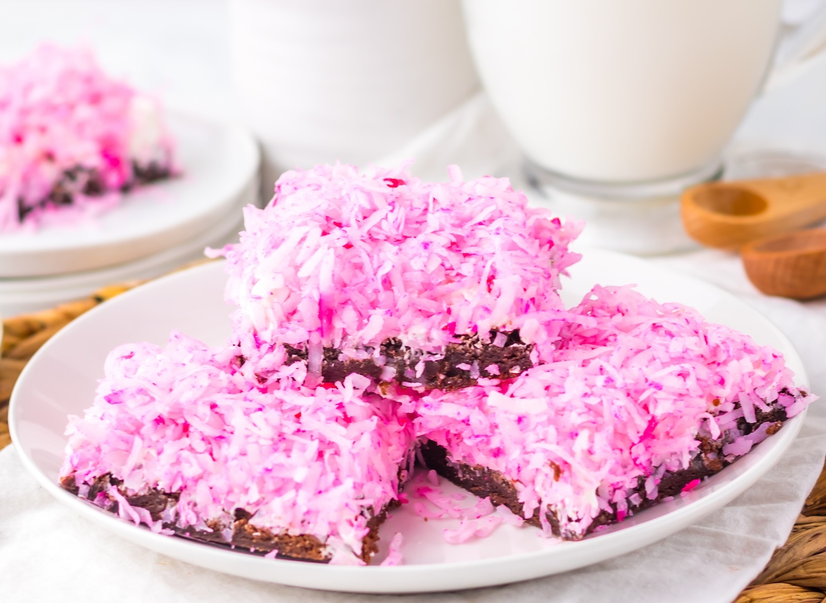 A plate of pink coconut-topped dessert bars stacked on a white plate, with a cup and another plate in the background.