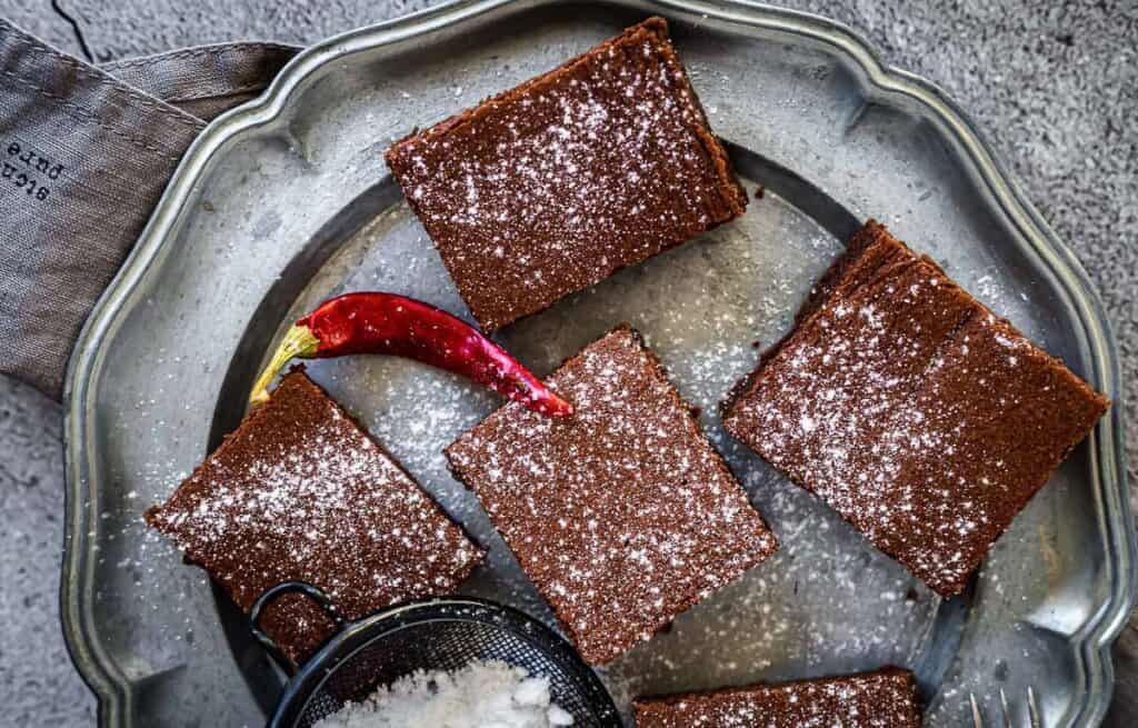 Four chocolate brownies on a round metal plate, garnished with a red chili pepper and powdered sugar. A small sieve with more sugar is nearby.