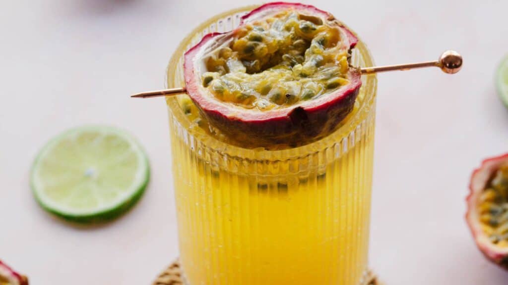 A glass of yellow juice garnished with half a passion fruit on a skewer. A slice of lime and a halved passion fruit are visible in the background.