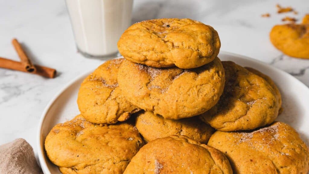 A plate of pumpkin cookies dusted with sugar, next to a glass of milk and cinnamon sticks on a marble surface.