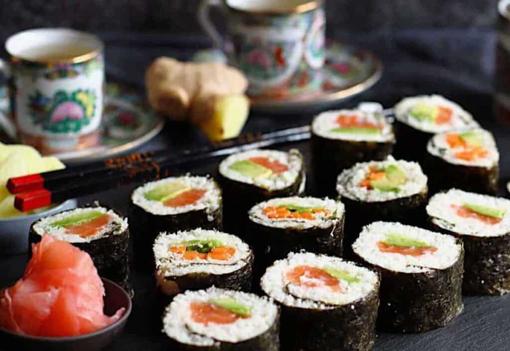 A platter of sushi rolls with seaweed wrapping, filled with vegetables and fish, is served next to pickled ginger, chopsticks, and decorative teacups.