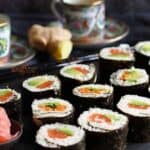 A platter of sushi rolls with seaweed wrapping, filled with vegetables and fish, is served next to pickled ginger, chopsticks, and decorative teacups.