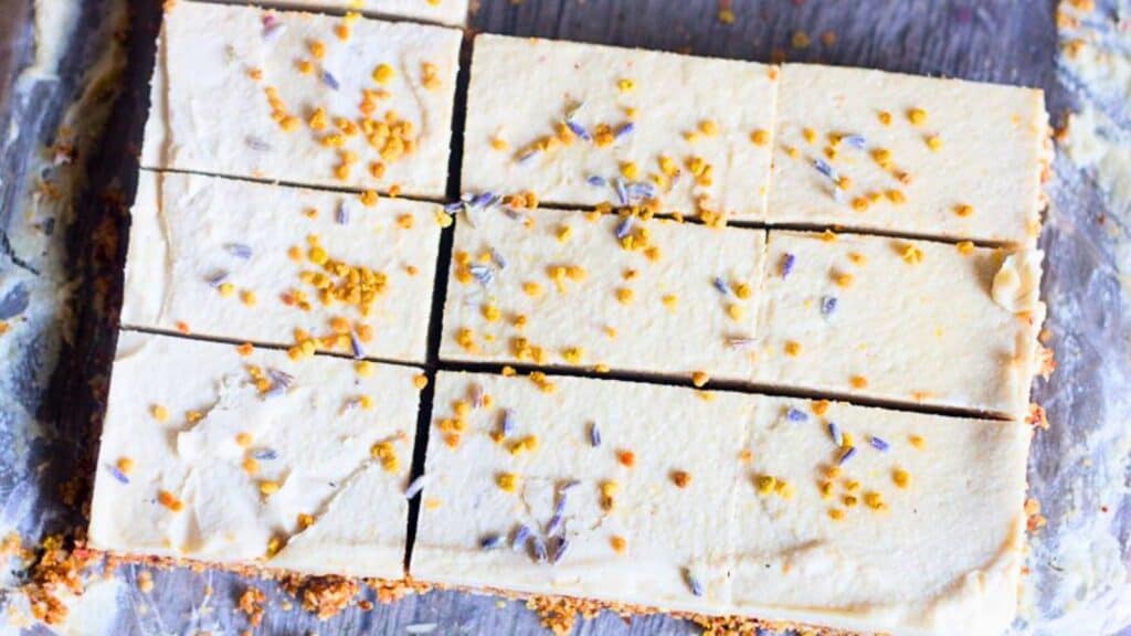 A tray of sliced cheesecake bars topped with lavender and crushed nuts, placed on a wooden surface.