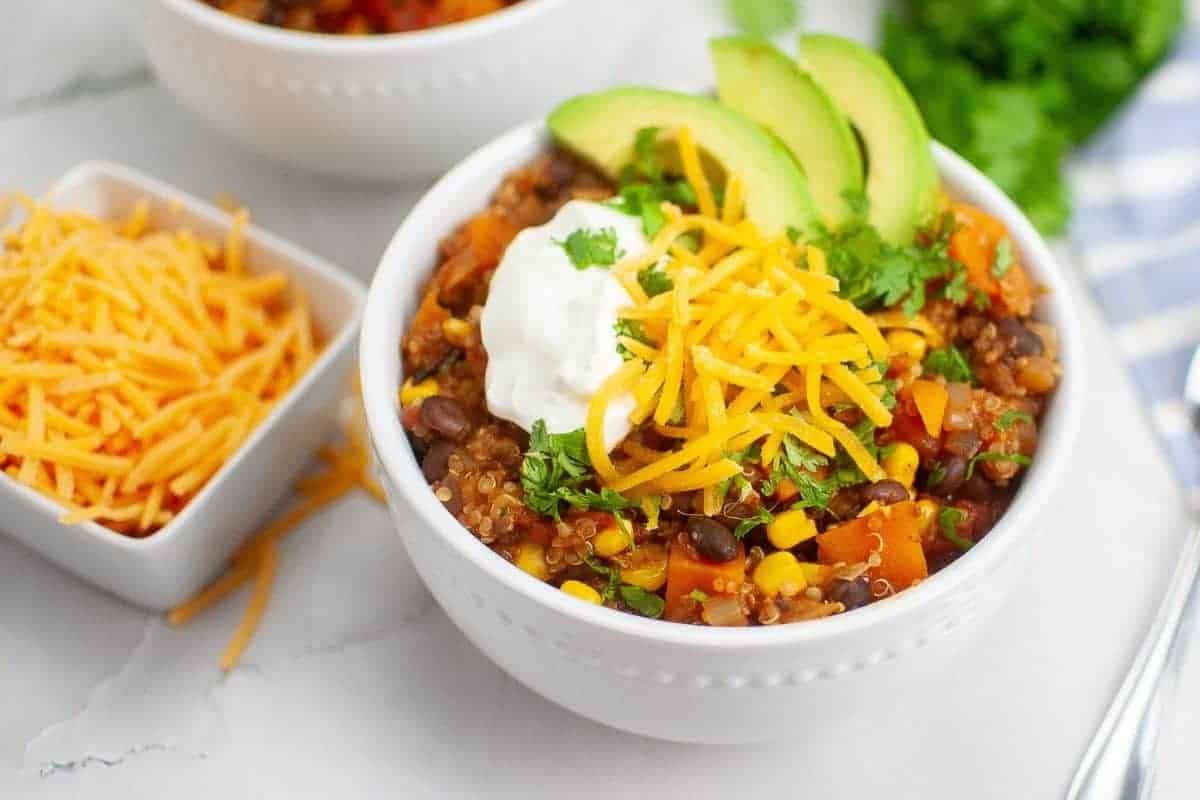 Bowl of sweet potato chili with quinoa topped with shredded cheese, sliced avocado, chopped cilantro and sour cream.