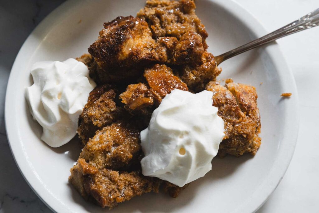 Plate of bread pudding with whipped cream, served with a spoon.