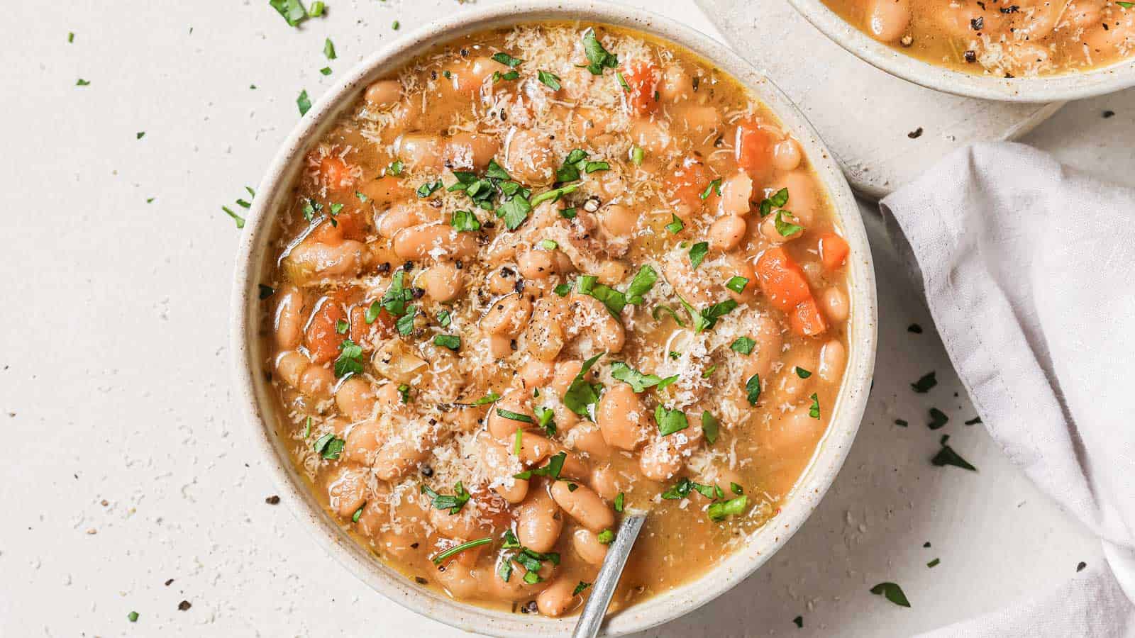 A bowl of bean stew with carrots, topped with grated cheese and chopped herbs, on a light surface.