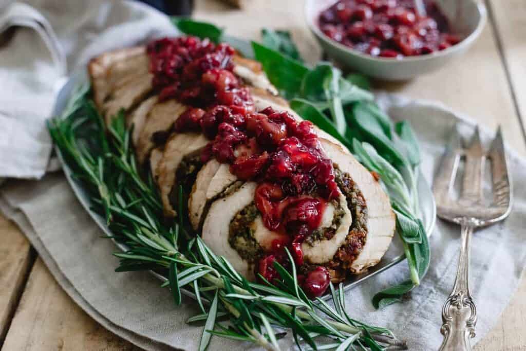 Sliced stuffed turkey breast topped with cranberry sauce, garnished with rosemary and sage leaves, on a white platter with a silver serving fork beside it.