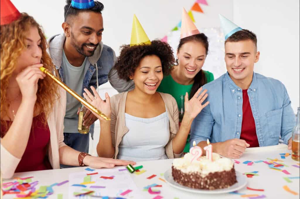 Five people celebrate a birthday with a cake decorated with the number 21, wearing party hats and surrounded by confetti.