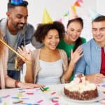 Five people celebrate a birthday with a cake decorated with the number 21, wearing party hats and surrounded by confetti.