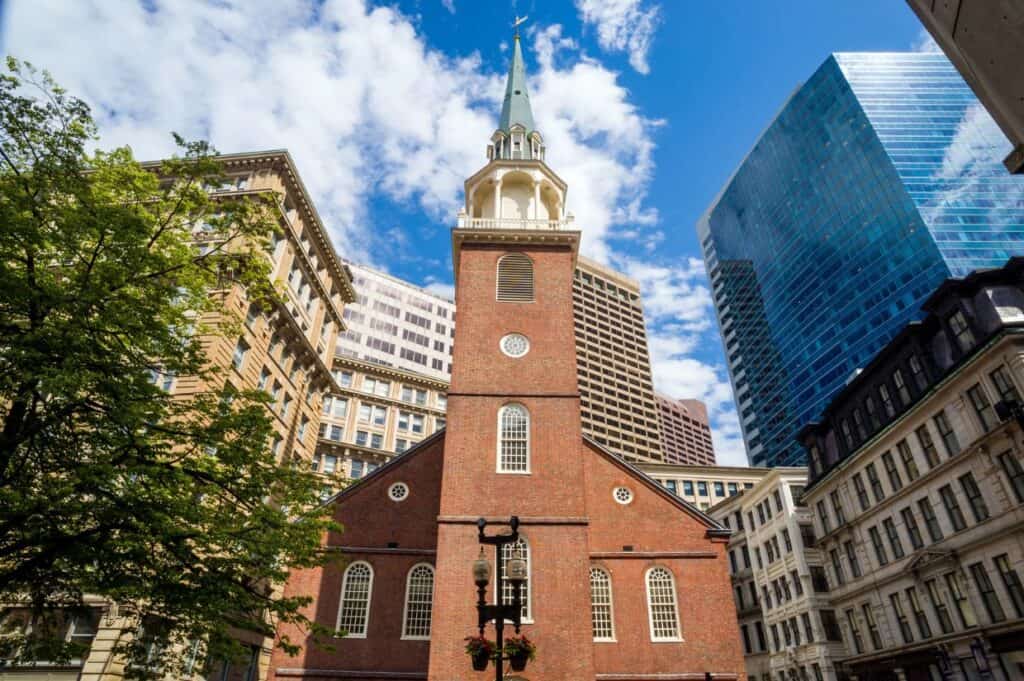 Discover this historic brick church, with its tall spire reaching towards the sky, nestled among modern high-rises and lush trees. A highlight for those on walking tours in Boston, it stands proudly beneath a partly cloudy sky.