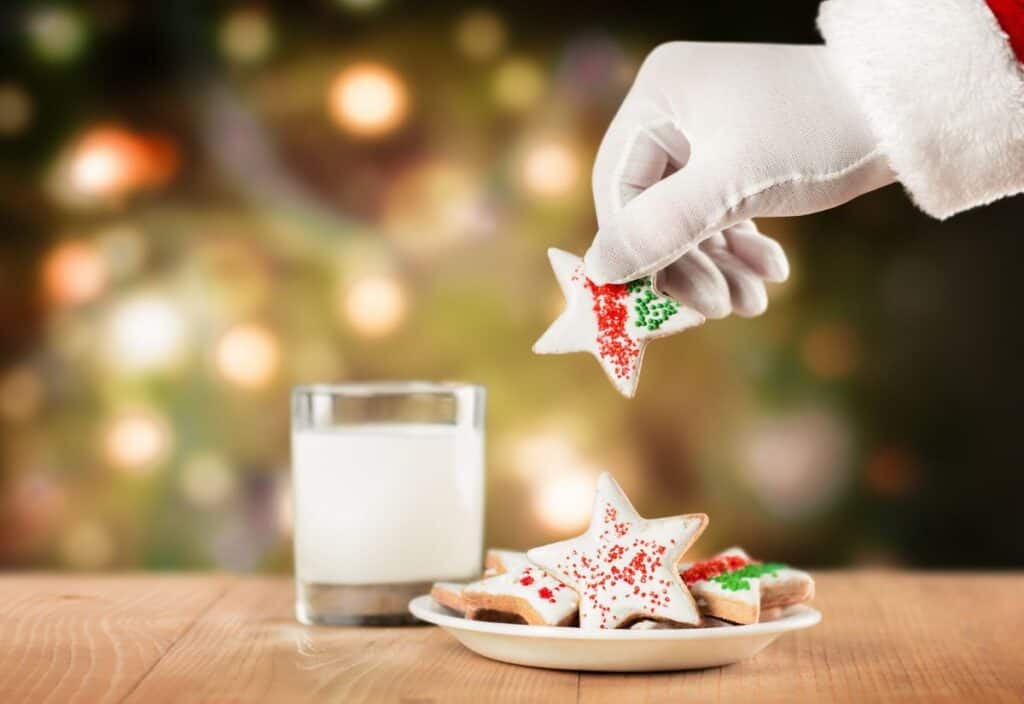 A gloved hand picks up a star-shaped cookie from a plate next to a glass of milk, with a blurred festive background.