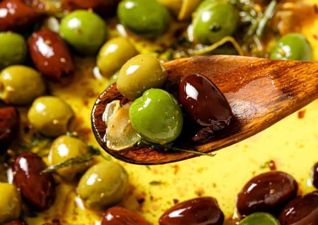 A wooden spoon holds a mix of green and black olives in oil, surrounded by more olives and herbs on a yellow surface.