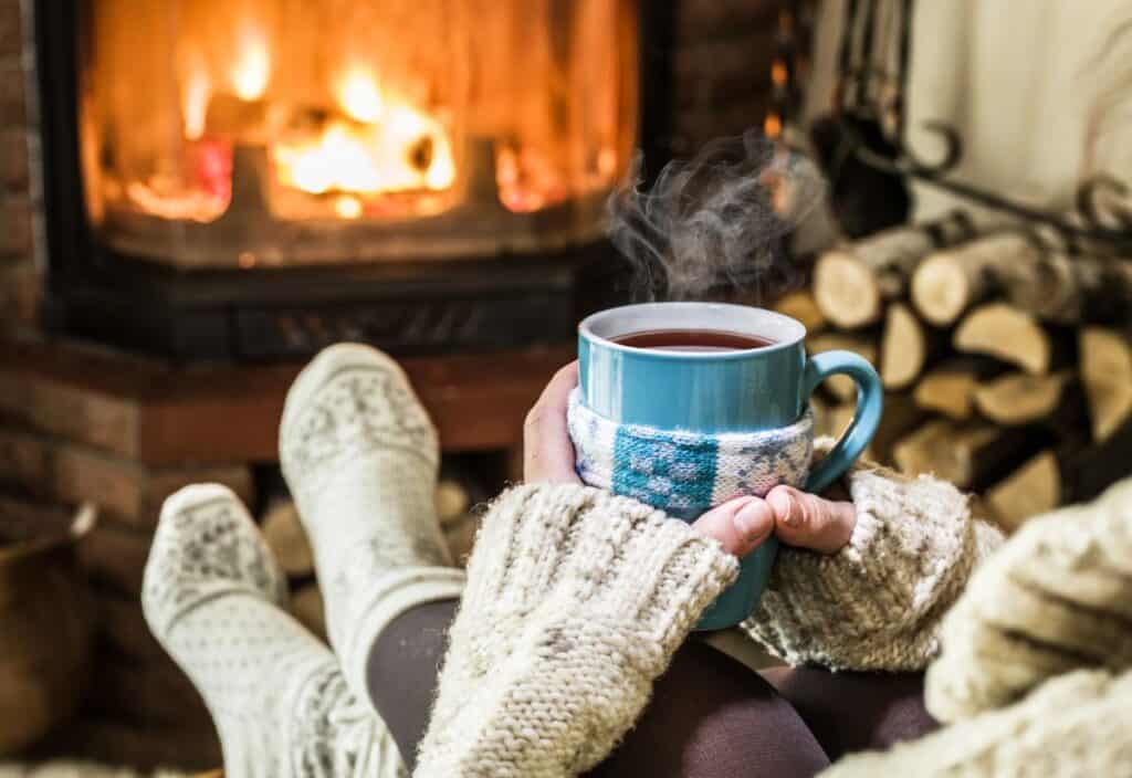 A person wearing cozy socks and a sweater embraces the essence of holiday hygge, holding a steaming mug near a lit fireplace with stacked firewood beside it.