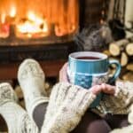 A person wearing cozy socks and a sweater embraces the essence of holiday hygge, holding a steaming mug near a lit fireplace with stacked firewood beside it.