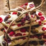 Stack of homemade white chocolate bark with dried raspberries, cranberries, and nuts, tied with twine, on a rustic wooden surface.