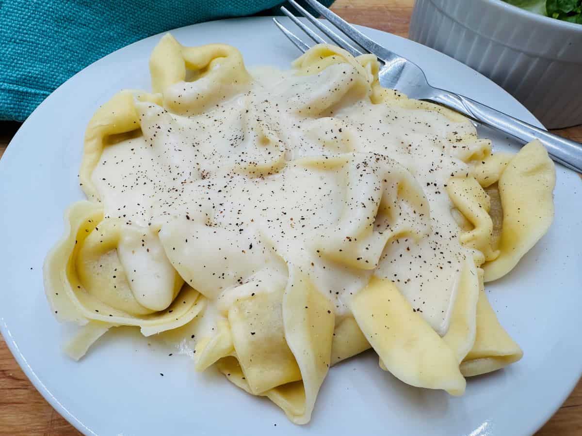 A plate of tortellini covered in a creamy white sauce, garnished with black pepper, next to a fork and a small bowl.