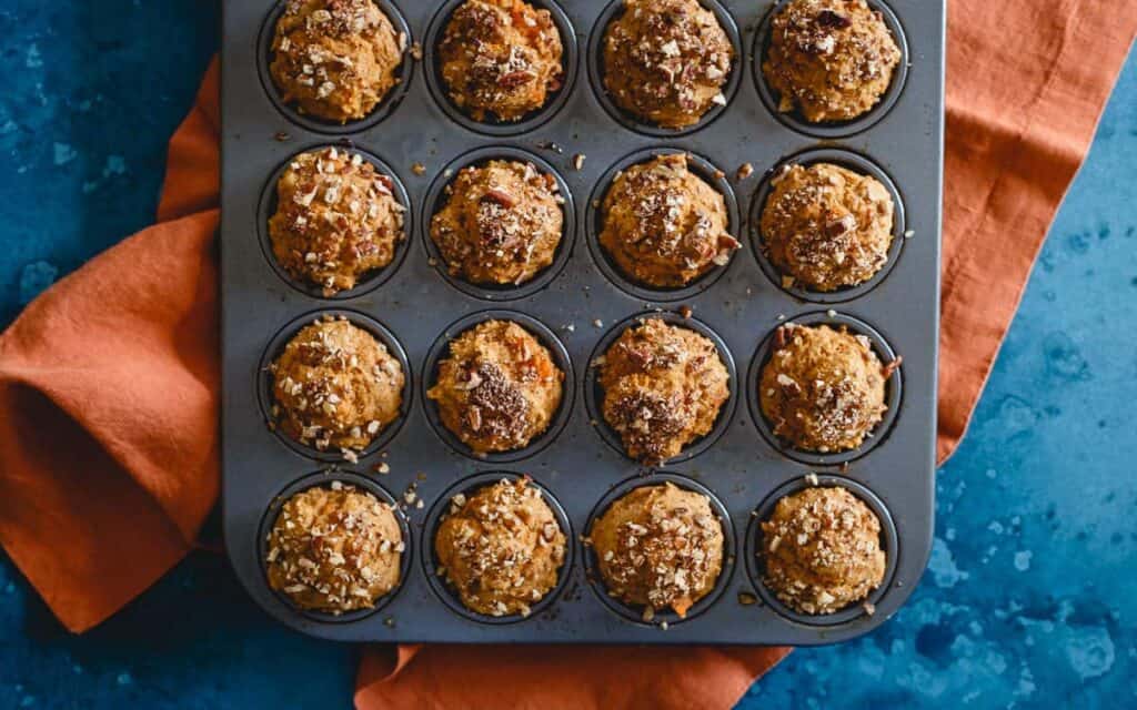 A muffin tray filled with 15 baked muffins, topped with nuts, on an orange cloth and blue surface.