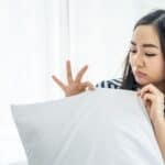 A woman examines a white pillow suspiciously, holding it with her left hand. She wears a striped shirt, with a neutral expression, against a light background.