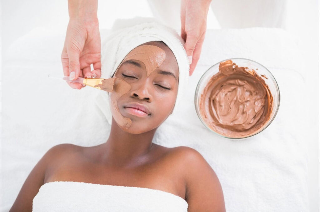 A person receives a facial treatment with a brown clay mask applied with a brush.