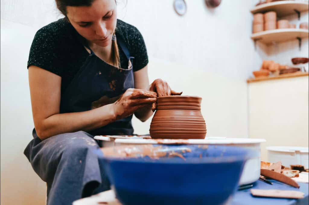 A person shaping a clay pot on a pottery wheel, surrounded by tools and materials in a workshop.