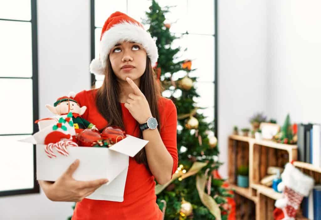 A woman in a Santa hat holds a box of Christmas decorations, looking thoughtful. A decorated Christmas tree is in the background.