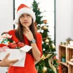 A woman in a Santa hat holds a box of Christmas decorations, looking thoughtful. A decorated Christmas tree is in the background.