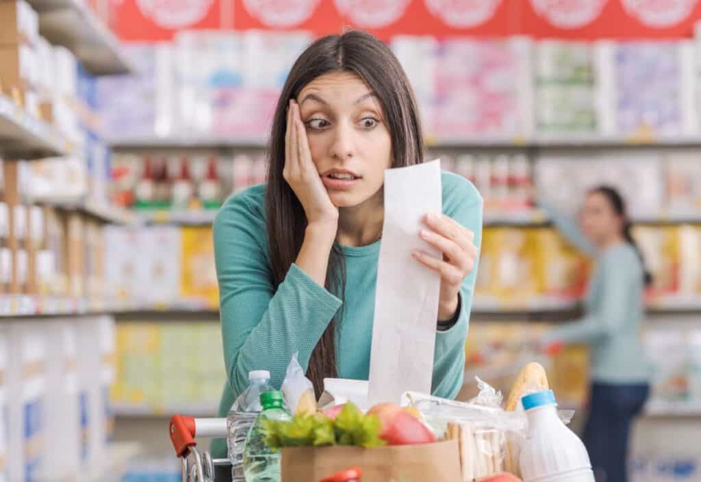 A woman looks surprised while holding a long receipt in a grocery store aisle, despite her shopping cart being full of low-cost foods.