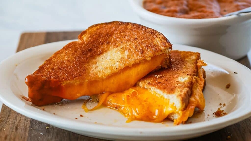 Two halves of a grilled cheese sandwich on a white plate, with melted cheese oozing out. A bowl of tomato soup is seen in the background.