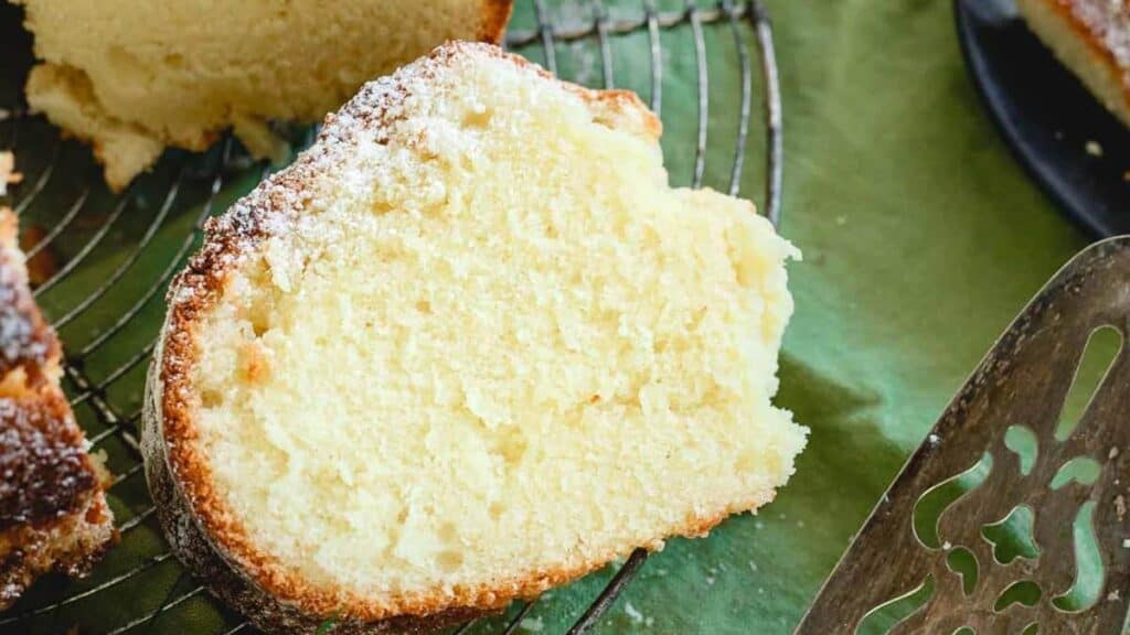 A slice of yellow cake dusted with powdered sugar on a wire rack, accompanied by a decorative cake server, on a green surface.