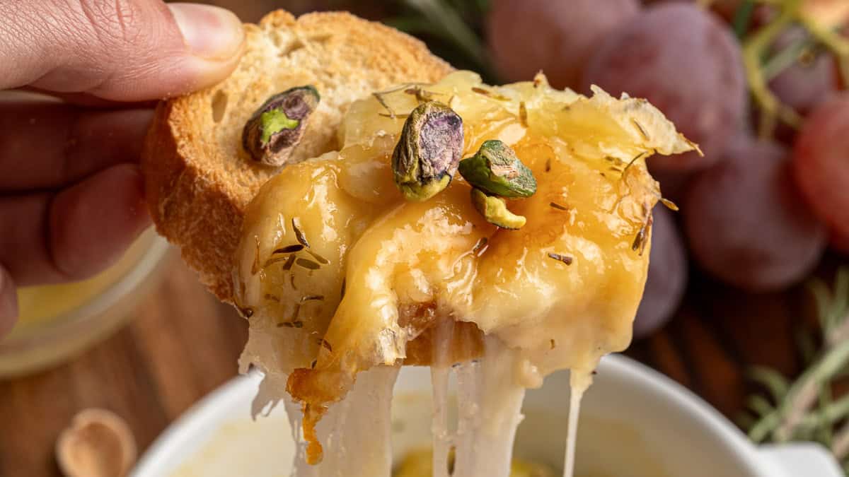 A hand holds a piece of bread topped with melted cheese and pistachios.