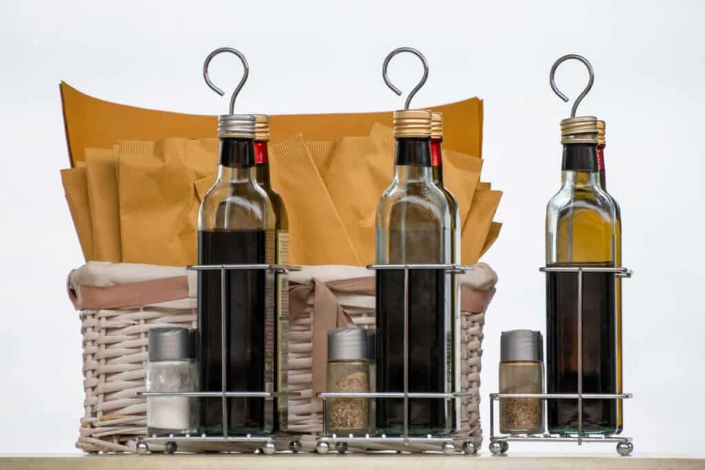 Three bottles of olive oil in metal holders, accompanied by salt and pepper shakers, in front of a wicker basket with paper bags.