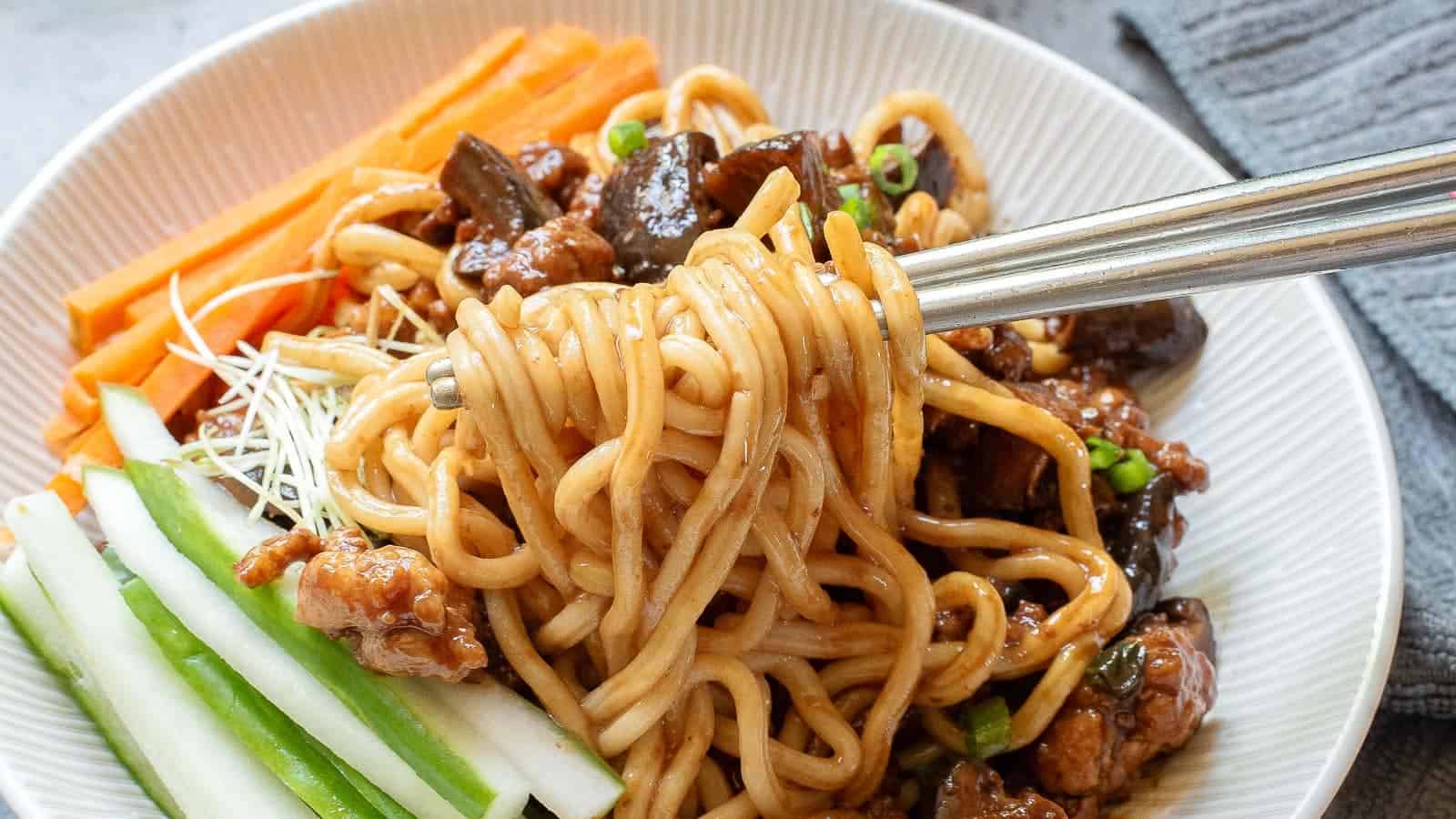 A bowl of noodles with sauce, garnished with sliced cucumbers, carrots, and bean sprouts. Using chopsticks, someone is lifting a portion of noodles. The dish is served in a white bowl, set on a light-colored table with a textured napkin nearby.