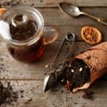 A glass teapot filled with tea sits on a wooden surface. Dried tea leaves spill from a paper bag, surrounded by dried citrus slices, a spoon, and a metal scoop.