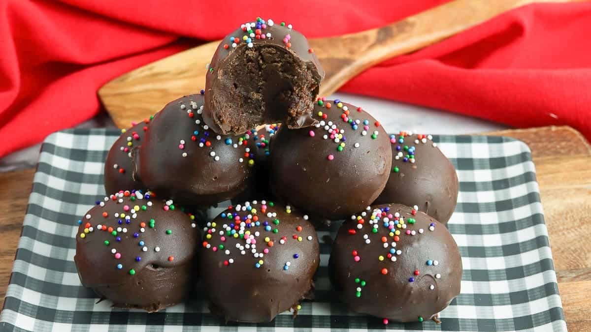 A stack of chocolate-covered cake balls topped with colorful sprinkles on a black and white checkered plate.