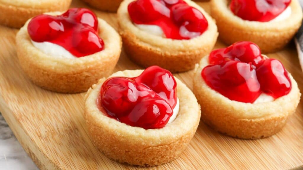 Mini cherry cheesecakes on a wooden board, each with a graham cracker crust and topped with glossy cherry filling.