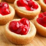 Mini cherry cheesecakes on a wooden board, each with a graham cracker crust and topped with glossy cherry filling.