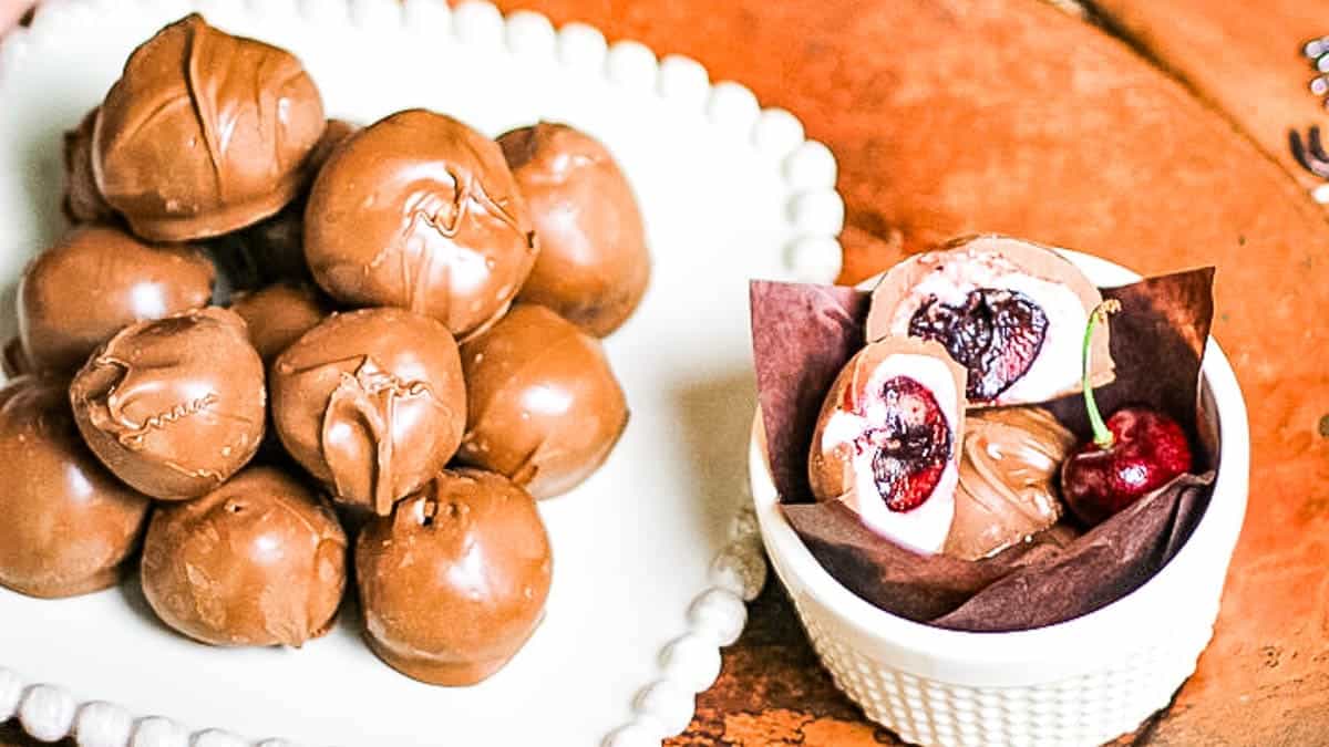 A plate of chocolate-covered cherries next to a bowl containing a cut open chocolate-covered cherry revealing the inside filling, placed on a wooden surface.