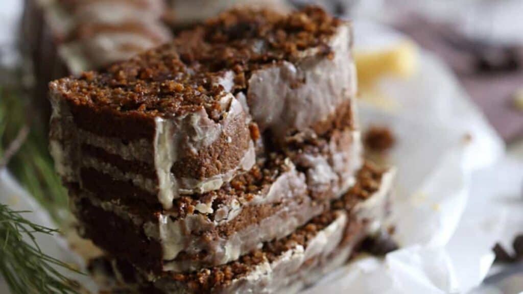 Close-up of a stack of sliced, frosted gingerbread loaf on a white plate, with a blurred background including greenery.