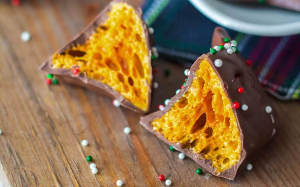 Two pieces of chocolate-coated honeycomb candy with colorful sprinkles are placed on a wooden surface.