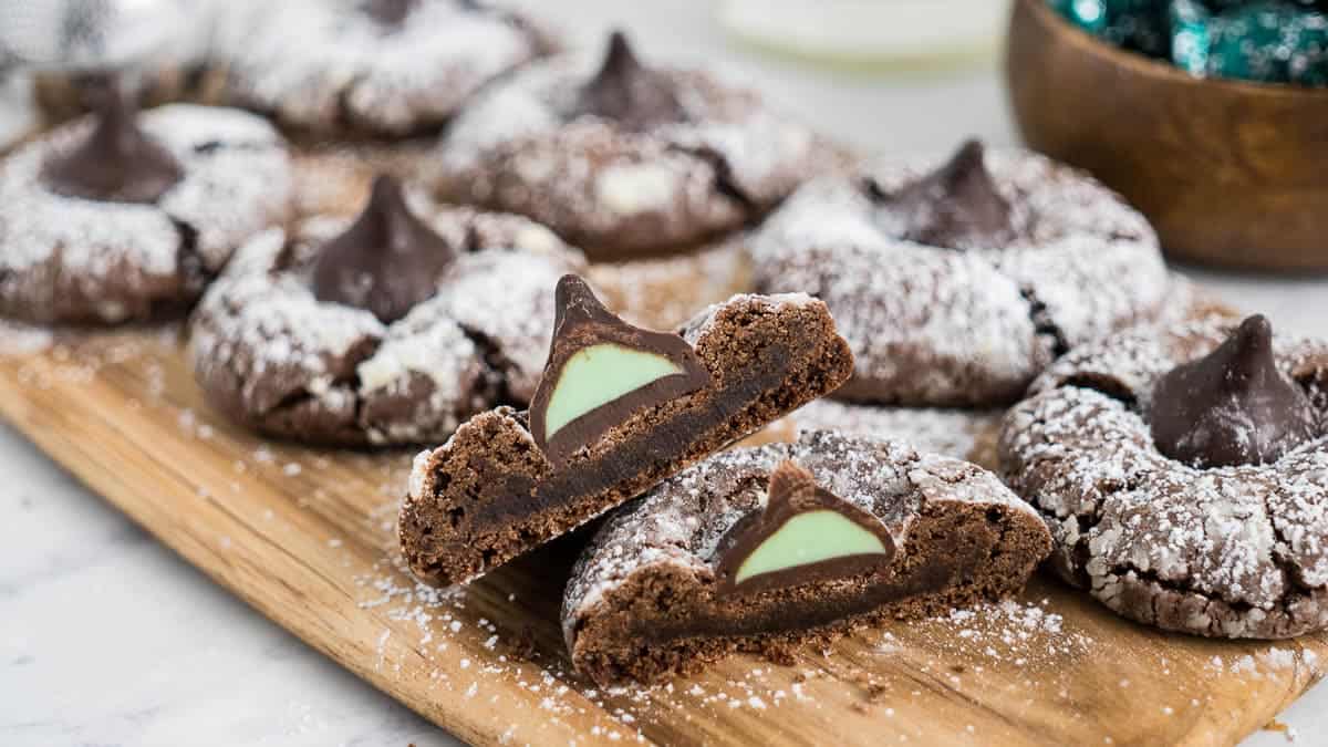 Close-up of chocolate cookies dusted with powdered sugar; one cookie is halved, revealing a mint filling and a chocolate kiss on top. 