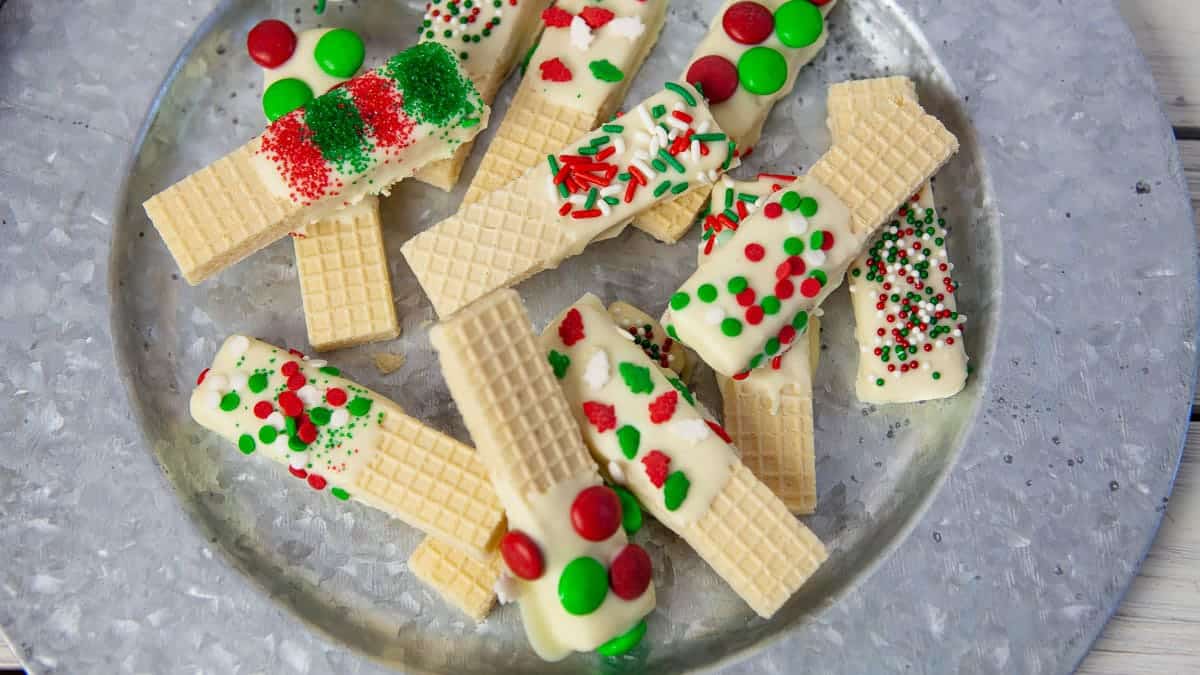 A metal plate with vanilla wafer cookies partially dipped in white chocolate and decorated with red, green, and white sprinkles and small candies.