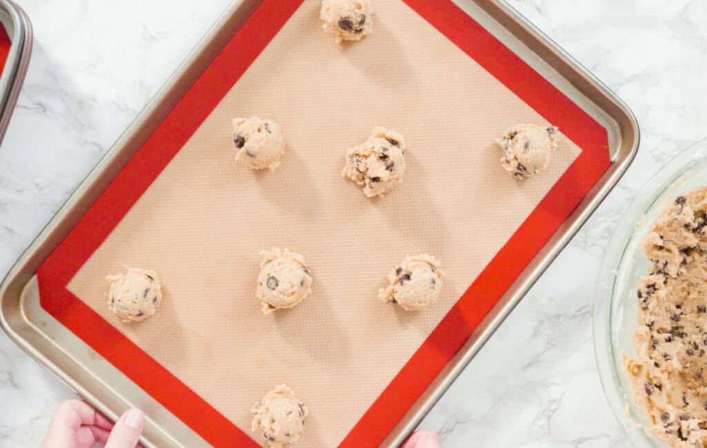 A baking sheet with a silicone mat holds nine scoops of chocolate chip cookie dough. A bowl of extra dough is nearby.