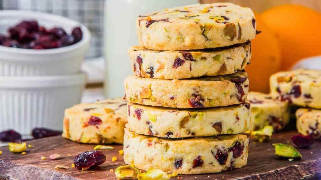 A stack of round cookies with visible pieces of pistachios and cranberries on a wooden surface, surrounded by scattered nuts and berries.