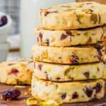 A stack of round cookies with visible pieces of pistachios and cranberries on a wooden surface, surrounded by scattered nuts and berries.