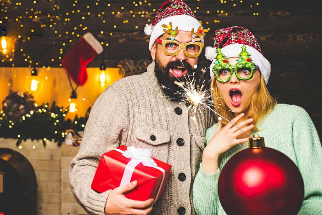 A man and woman wearing festive attire and glasses hold a sparkler, a red gift, and a large red ornament in a decorated room with holiday lights and stockings.