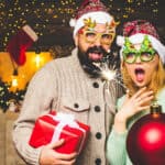 A man and woman wearing festive attire and glasses hold a sparkler, a red gift, and a large red ornament in a decorated room with holiday lights and stockings.