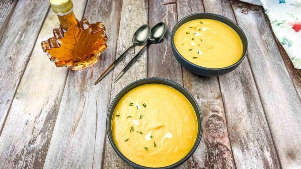 Two bowls of cream of butternut squash & sweet potato soup garnished with herbs on a wooden table.