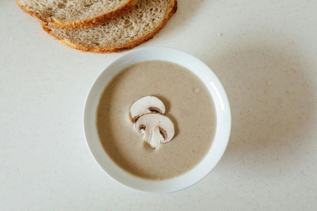 A bowl of mushroom soup with sliced mushrooms on top, placed on a light surface next to slices of bread.