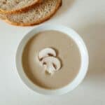 A bowl of mushroom soup with sliced mushrooms on top, placed on a light surface next to slices of bread.